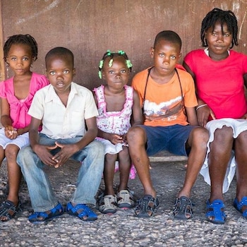 Cinco niños y jóvenes están sentados en fila sobre un banco con camisetas de varios colores y sandalias