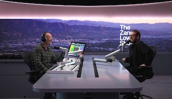 Jonah Hill y un hombre en un estudio de radio con micrófonos. Ambos sentados. Una pantalla detrás de ellos muestra montañas y una ciudad