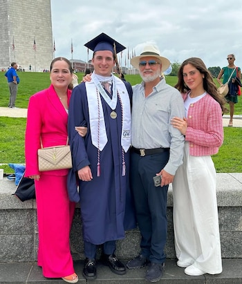 Cuatro personas posan para una foto al aire libre: un joven con toga y birrete azul, estola blanca, y tres adultos. Al fondo, el Monumento a Washington