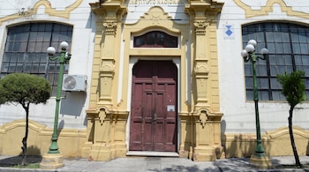 Fachada de un edificio con cartel de 'INSTITUTO NACIONAL', una puerta de madera, dos ventanas arqueadas, dos faroles verdes y dos árboles pequeños.