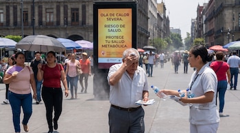 Vista de calle concurrida en la Ciudad de México bajo el sol; un hombre mayor se refresca mientras una mujer le ofrece agua y folletos junto a una valla publicitaria.