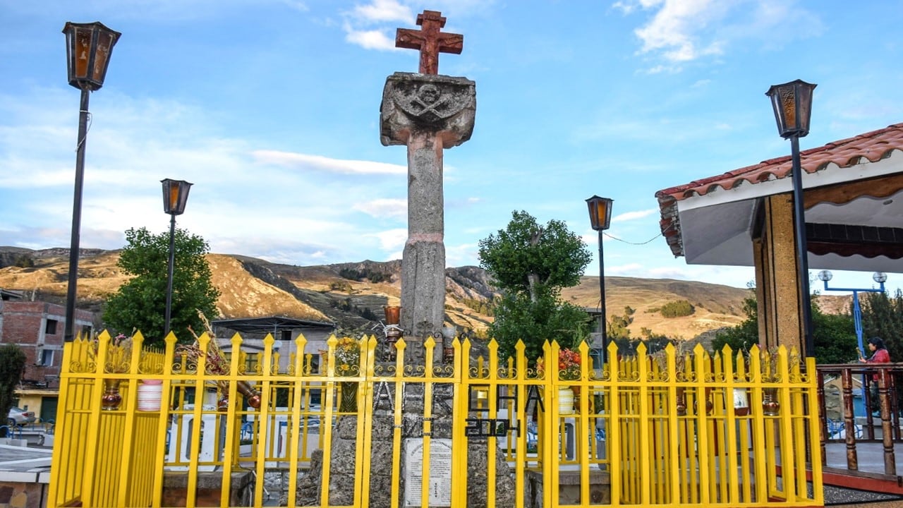 Un monumento con cruz de piedra y una cerca amarilla se alza frente a un paisaje montañoso con construcciones.