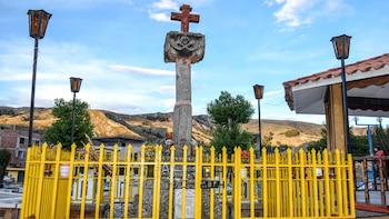 Monumento con cruz de piedra, valla amarilla, farolas de estilo antiguo, árboles, construcciones y montañas bajo un cielo azul nublado