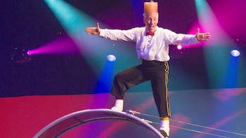 Un hombre de circo con camisa blanca, pajarita roja y pantalones negros se equilibra sobre una rueda metálica, con el pelo rubio y levantado