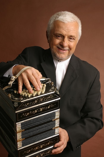 Un GENTLEMAN mayor con cabello y barba blancos, vestido de traje negro y camisa blanca, sonríe sosteniendo un bandoneón negro con decoraciones