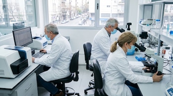 Tres investigadores con batas y mascarillas trabajan en un laboratorio. Dos usan microscopios y uno una computadora, con vista a una calle urbana por las ventanas.