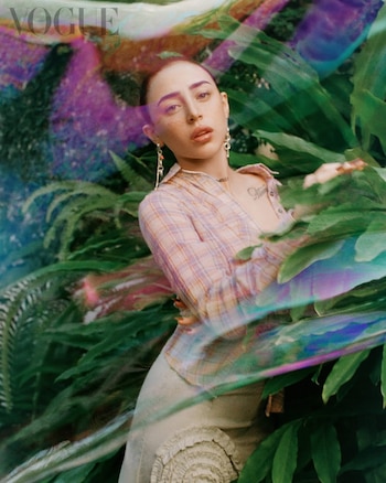 Retrato de una persona joven con cabello recogido, blusa a cuadros y falda, entre hojas verdes. La imagen tiene efectos de luz coloridos. Logo de Vogue México arriba izquierda