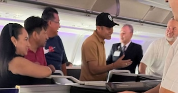 Un hombre con gorra negra y polo marrón sonríe y gesticula con las manos en un avión, rodeado de otras cinco personas que también sonríen en la cabina