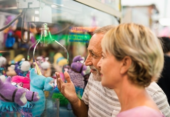 Un hombre y una mujer de perfil observan los peluches dentro de una máquina de gancho con una garra metálica. Se aprecian unicornios azules y otros muñecos de colores