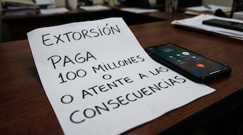 Un papel sobre un escritorio con el texto escrito EXTORSIÓN PAGA 100 MILLONES O ATENTE A LAS CONSECUENCIAS junto a un teléfono inteligente.