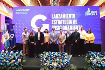Diez personas posan en un escenario ante una pantalla azul de AGEXPORT, con arreglos florales en el frente y la bandera de Guatemala a un lado