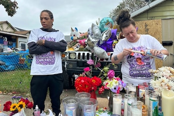 Un hombre con los brazos cruzados y una mujer que enciende una vela están junto a un altar con flores, velones y globos frente a un vehículo