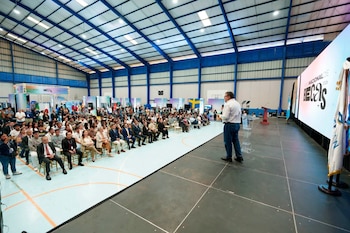 Un hombre con micrófono habla en un escenario ante personas sentadas en un pabellón techado junto a una pantalla gigante