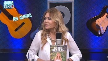 Mujer rubia sentada con un micrófono, sostiene un libro titulado "El Poder de Ser Tú" en un estudio de televisión con guitarras y carteles