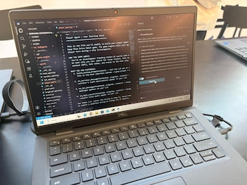 Una laptop Dell gris abierta sobre una mesa oscura. La pantalla muestra código Python en un IDE y el teclado está en primer plano. Un cable negro se conecta al lateral