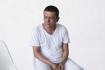 Hombre de mediana edad con camiseta y pantalón blancos, sentado en una silla blanca. Sus manos están esposadas y su mirada se dirige a la izquierda.
