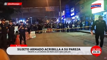 Policías y una multitud de personas están presentes en una calle oscura, con una cinta de seguridad amarilla bloqueando el paso, frente a tiendas iluminadas