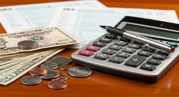 Primer plano de una calculadora financiera con un bolígrafo sobre ella, billetes de dólares y monedas al lado, y formularios de impuestos desenfocados en un escritorio de madera.