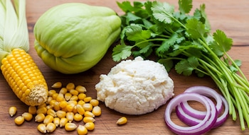 Un chayote verde, un elote, requesón blanco, un manojo de cilantro fresco y rodajas de cebolla morada se encuentran sobre una tabla de madera marrón.
