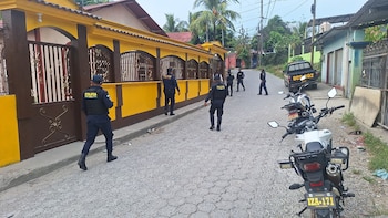 Varios agentes de la Policía Nacional Civil de Guatemala caminan por una calle adoquinada frente a casas, con un camión y motocicletas estacionados