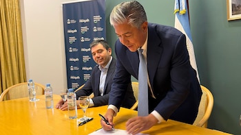 Dos hombres en traje en una mesa de madera. Uno, de cabello gris, firma un documento con un bolígrafo. El otro, sentado, sonríe. Hay botellas de agua y vasos
