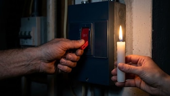 Una mano acciona un interruptor rojo en un panel eléctrico oscuro, mientras otra mano sostiene una vela blanca encendida