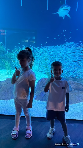 Niña y niño posan frente a un acuario. Ambos tienen pulgares arriba. El acuario contiene un tiburón, muchos peces, rocas y plantas acuáticas