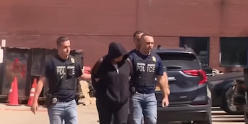 Dos agentes de policía escoltan a una persona encapuchada y esposada. Se observa un vehículo, un edificio de ladrillo, conos y un contenedor