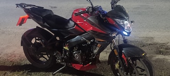 Una motocicleta deportiva roja y negra con detalles en gris oscuro, con sus luces delanteras y traseras encendidas, estacionada sobre tierra y pasto