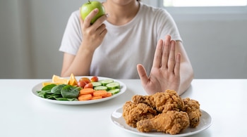 Una persona sentada en una mesa blanca, sosteniendo una manzana verde y mostrando la palma para rechazar pollo frito, con un plato de ensalada y frutas al fondo.