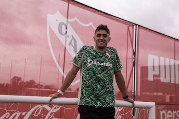 Hombre en camiseta verde con estampado de camuflaje, escudo C.A.I., logos Sportsbet y Puma. Apoyado en portería blanca. Fondo con valla roja y escudo C.A.I., logo Coca-Cola