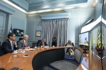 Personas sentadas alrededor de una mesa de reuniones semicircular en una sala azul, participando en una videoconferencia con el presidente Zelensky visible en pantalla