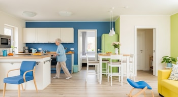 Vista de una cocina y sala de estar con una persona mayor caminando. Se observan barandas en la isla de la cocina, sillas ergonómicas, y un diseño luminoso.