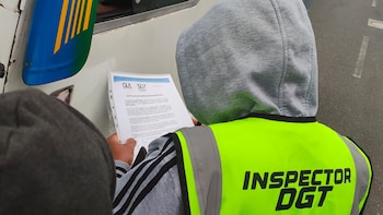 Vista trasera de un inspector con chaleco amarillo que dice 'INSPECTOR DGT' y una sudadera gris, sosteniendo un documento cerca de un vehículo