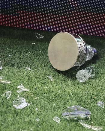 Un trofeo de plata y cristal roto yace en un campo de césped verde, rodeado de fragmentos de cristal. Al fondo, una pantalla LED con luces de colores