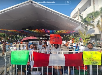 Entusiastas fans de Grupo 5 se reúnen en Oaxaca, México, tras una barrera. Sostienen una bandera peruana y carteles bajo una carpa decorada con papel picado