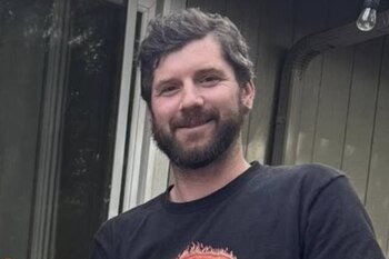 Retrato de un hombre de cabello castaño y barba que viste una camiseta oscura frente a la pared exterior de una casa