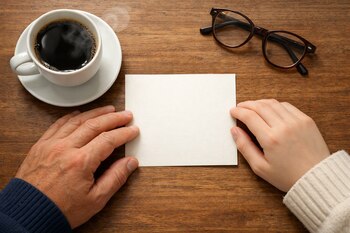 Vista cenital de dos manos sobre una mesa de madera, sosteniendo una tarjeta en blanco. Una taza de café humeante y unas gafas complementan la escena.