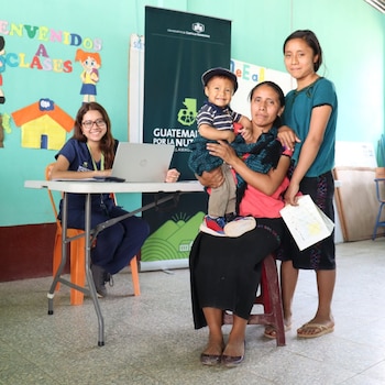 Una mujer sentada en una mesa con laptop, otra mujer con un bebé en brazos y una niña de pie. Un mural y un cartel de nutrición se ven de fondo