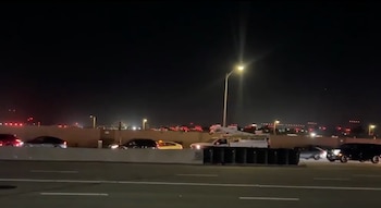 Vista nocturna de una carretera con coches transitando y un muro de contención, con las luces del aeropuerto de Los Ángeles visibles al fondo