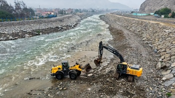 Río con orillas de roca, dos máquinas de construcción amarillas en el lecho seco, una excavadora y una cargadora frontal, edificios al fondo