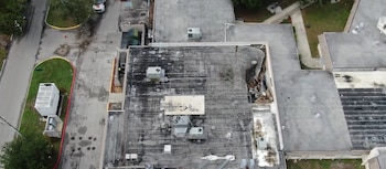 Vista aérea de un edificio con un colapso parcial de su techo plano, mostrando unidades de aire acondicionado, escombros, un vehículo y zonas verdes