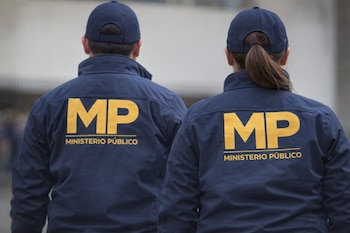Vista posterior de dos agentes, un hombre y una mujer, del Ministerio Público vestidos con chaquetas y gorras azul marino con el logo "MP" en amarillo.