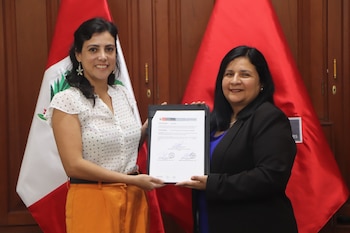 Dos mujeres sonrientes sostienen un documento firmado frente a dos banderas peruanas. La mujer de la izquierda viste blusa blanca y pantalón naranja, la de la derecha un blazer negro