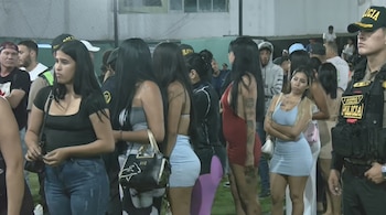 Varias personas permanecen de pie junto a agentes de policía uniformados con chalecos tácticos en una cancha con césped artificial.