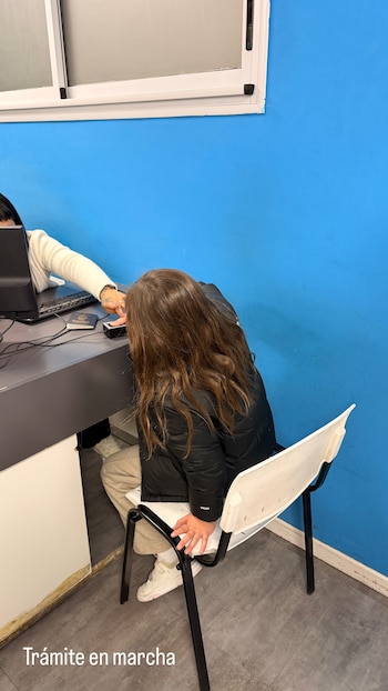 Niño sentado en silla blanca frente a escritorio gris. Adulto con brazo extendido hacia un lector de huellas. Pasaporte y monitor en el escritorio. Pared azul