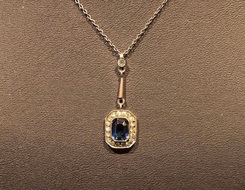 Collar de cadena metálica fina con un colgante. El colgante presenta una gema azul rectangular, rodeada por veinticuatro gemas claras. Fondo marrón texturizado