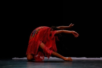 Un bailarín con un atuendo rojo con flecos y bordados azules se arrodilla en un escenario oscuro, con los brazos extendidos y el torso inclinado hacia adelante