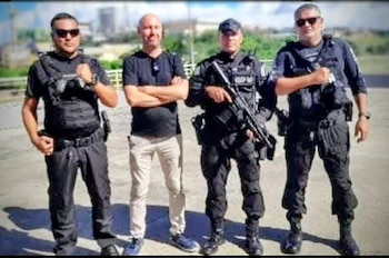 Cuatro hombres de pie. Tres visten uniformes tácticos oscuros con chalecos y gafas de sol, uno sostiene un rifle. Un cuarto viste ropa casual