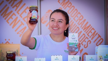 Mujer sonriente sujeta un frasco de miel y una barra de chocolate oscuro Kelfa frente a un cartel con letras naranjas